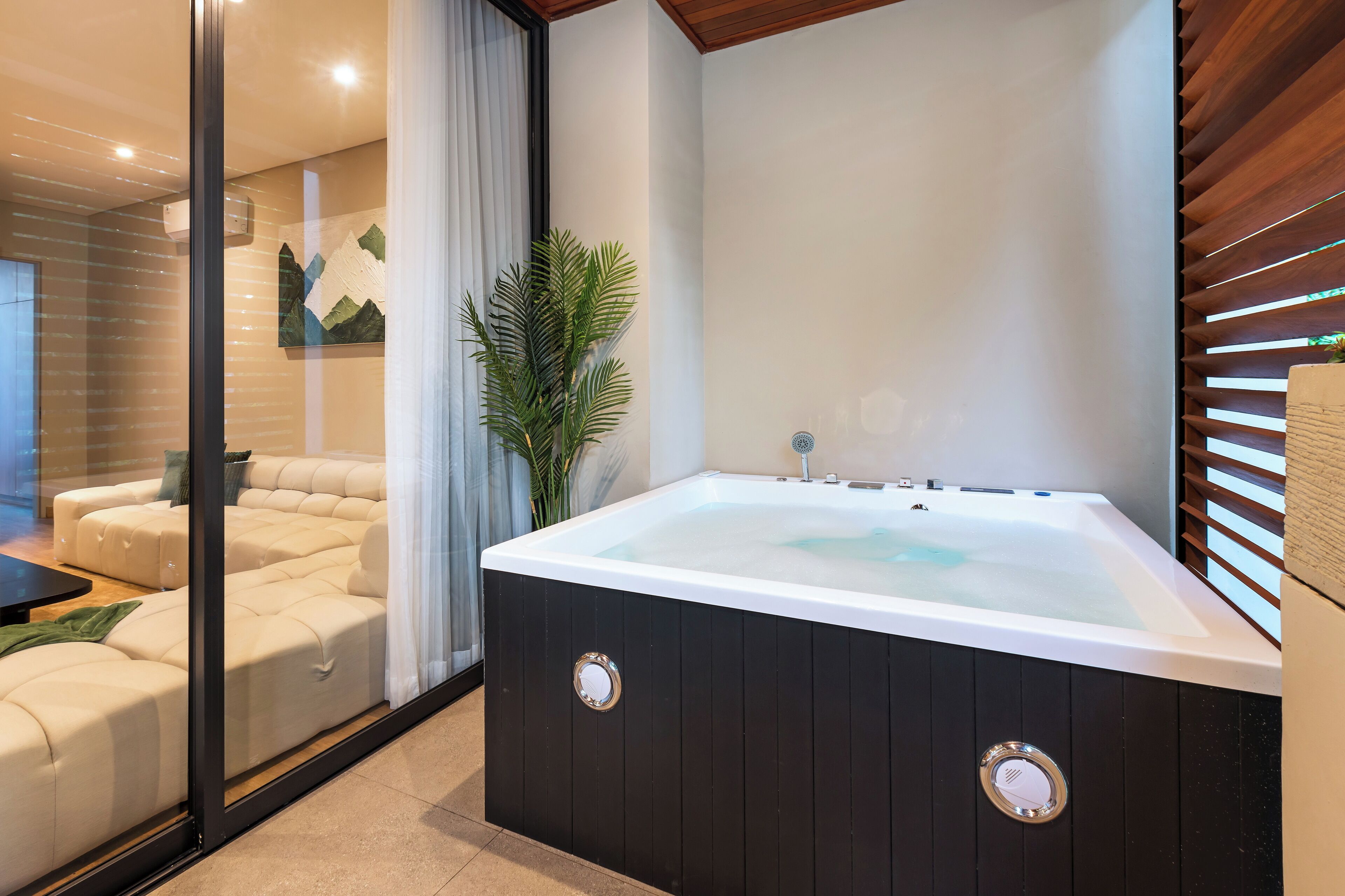 A modern bathroom with a large bathtub, a glass shower enclosure, and a seating area with a sofa and a potted plant.