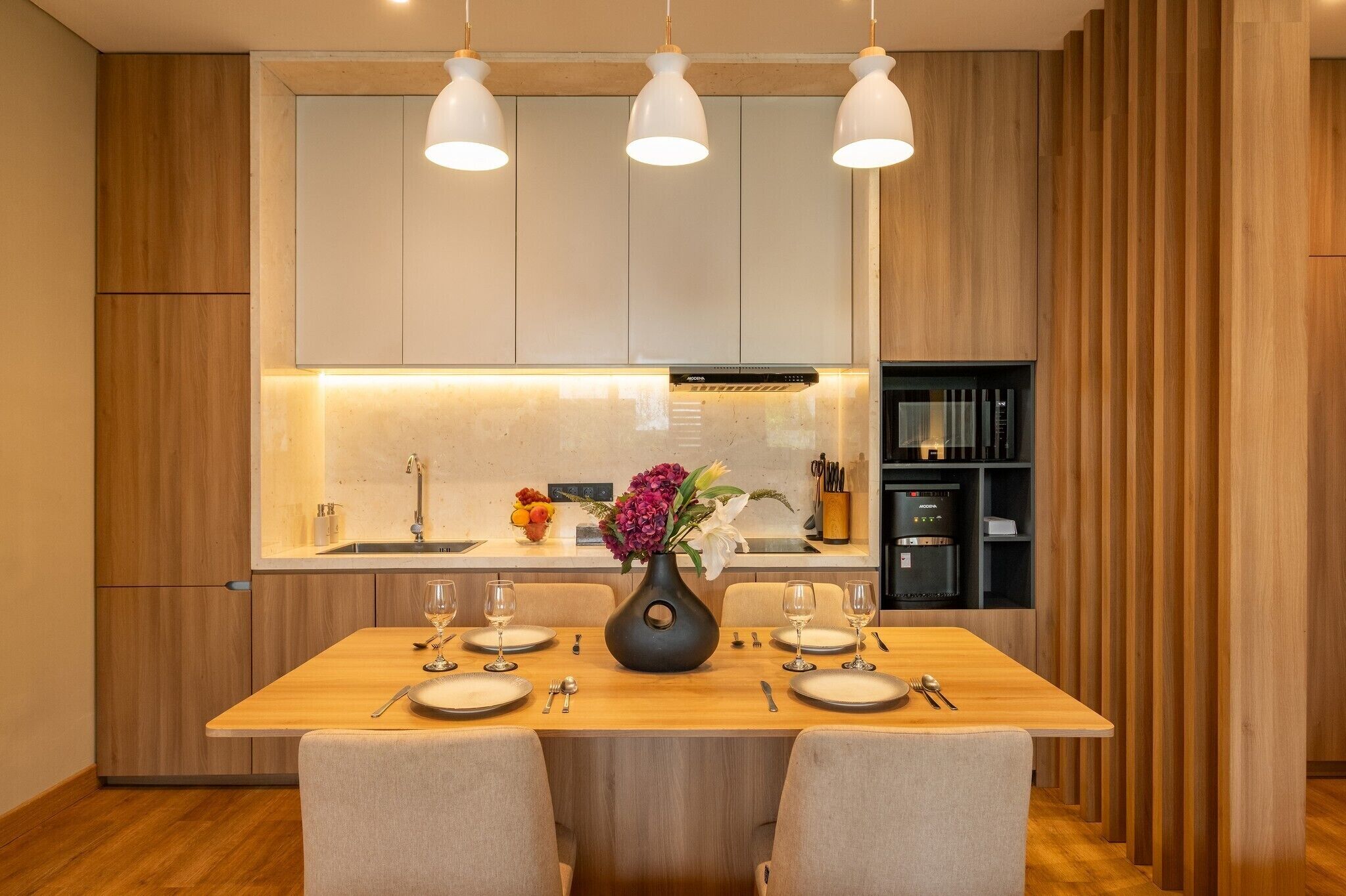A modern kitchen with wooden cabinets, a central island with a vase of flowers, and pendant lighting.