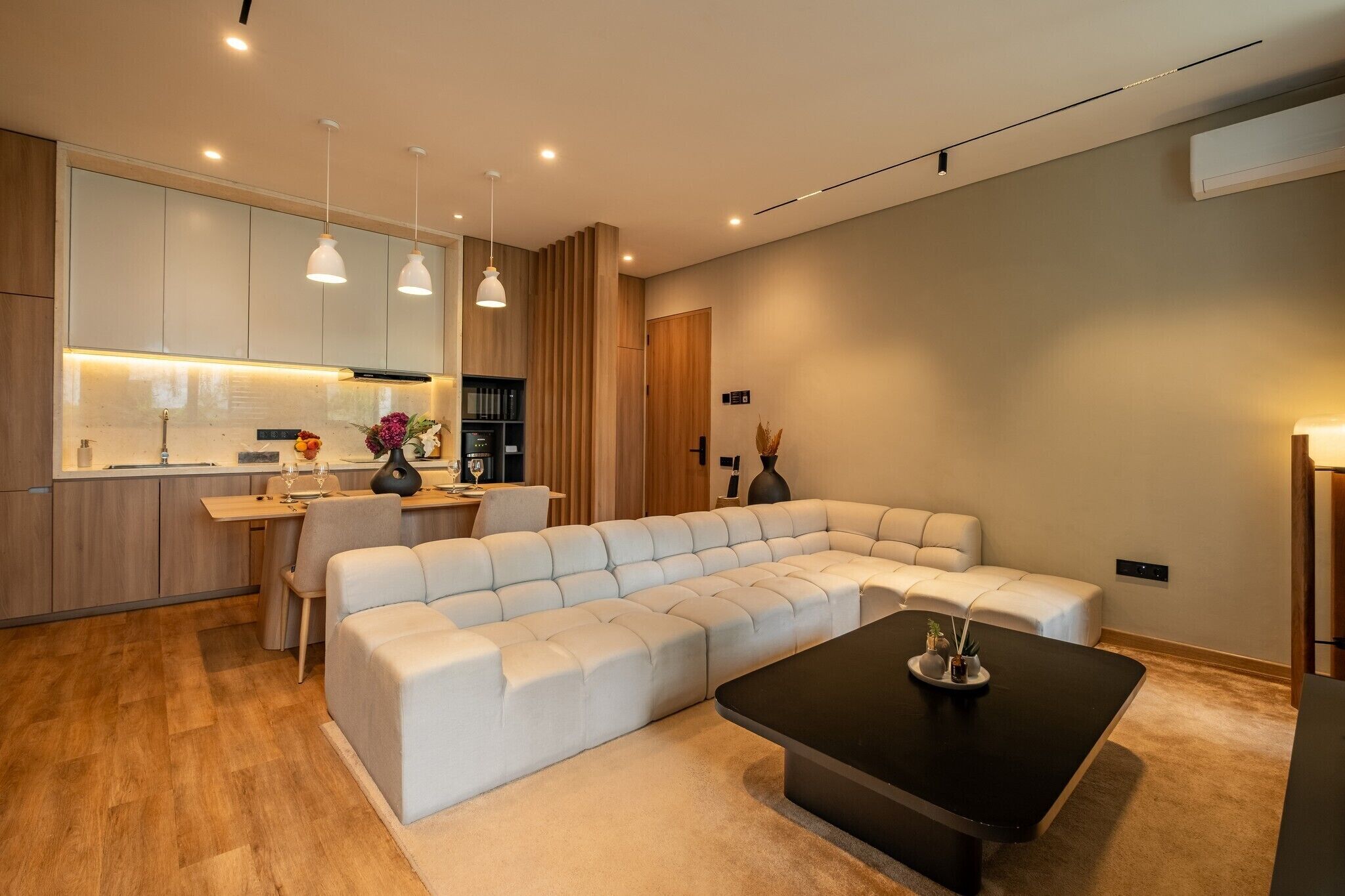 Ein modernes Wohnzimmer mit einer Eckcouch, einem Essbereich mit Tisch und Stühlen sowie einer Küche mit eingebauten Schränken und Pendelleuchten.