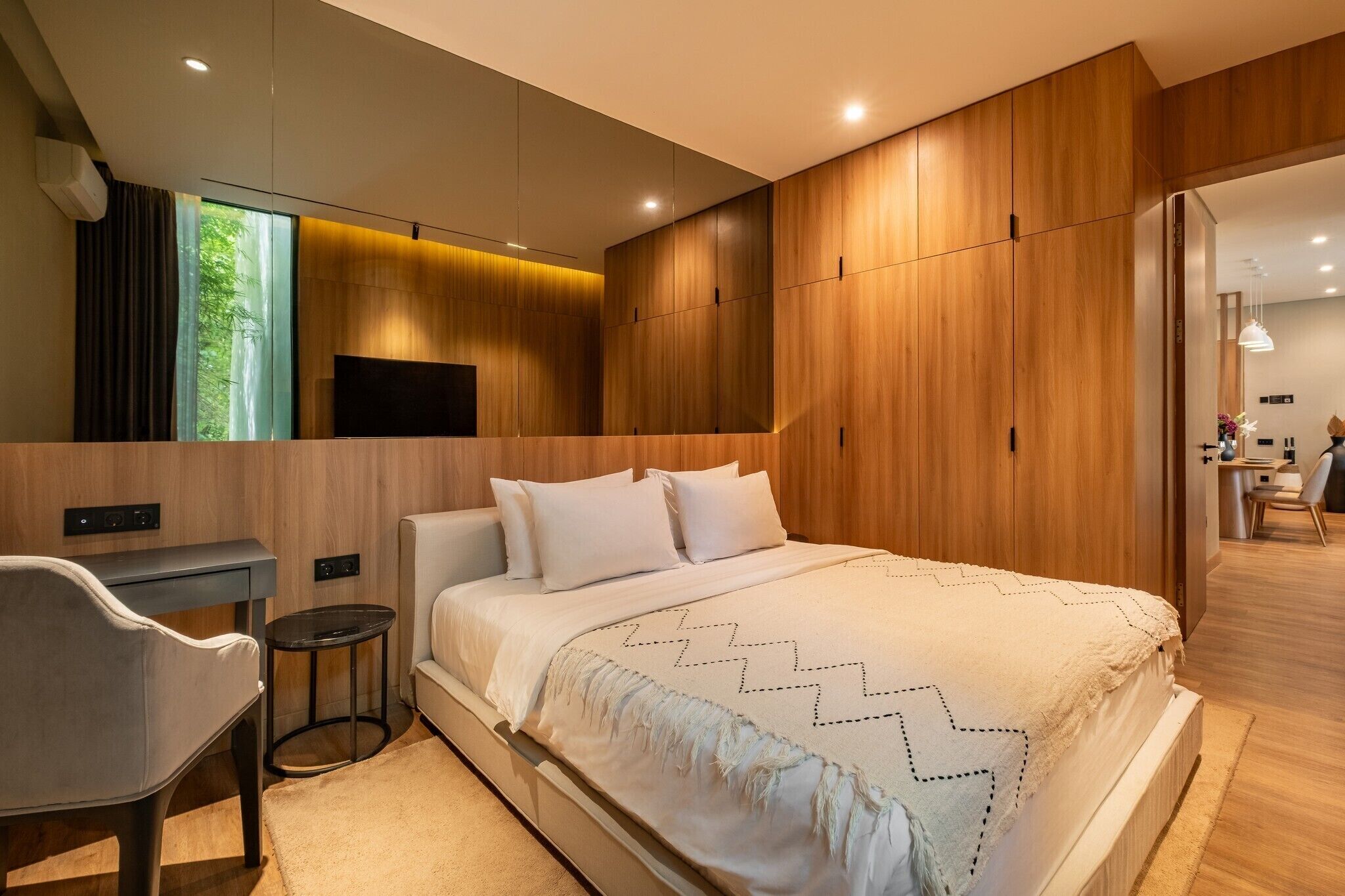 A modern bedroom with a large bed, a desk, and a chair. There is a TV mounted on the wall and a window with curtains.