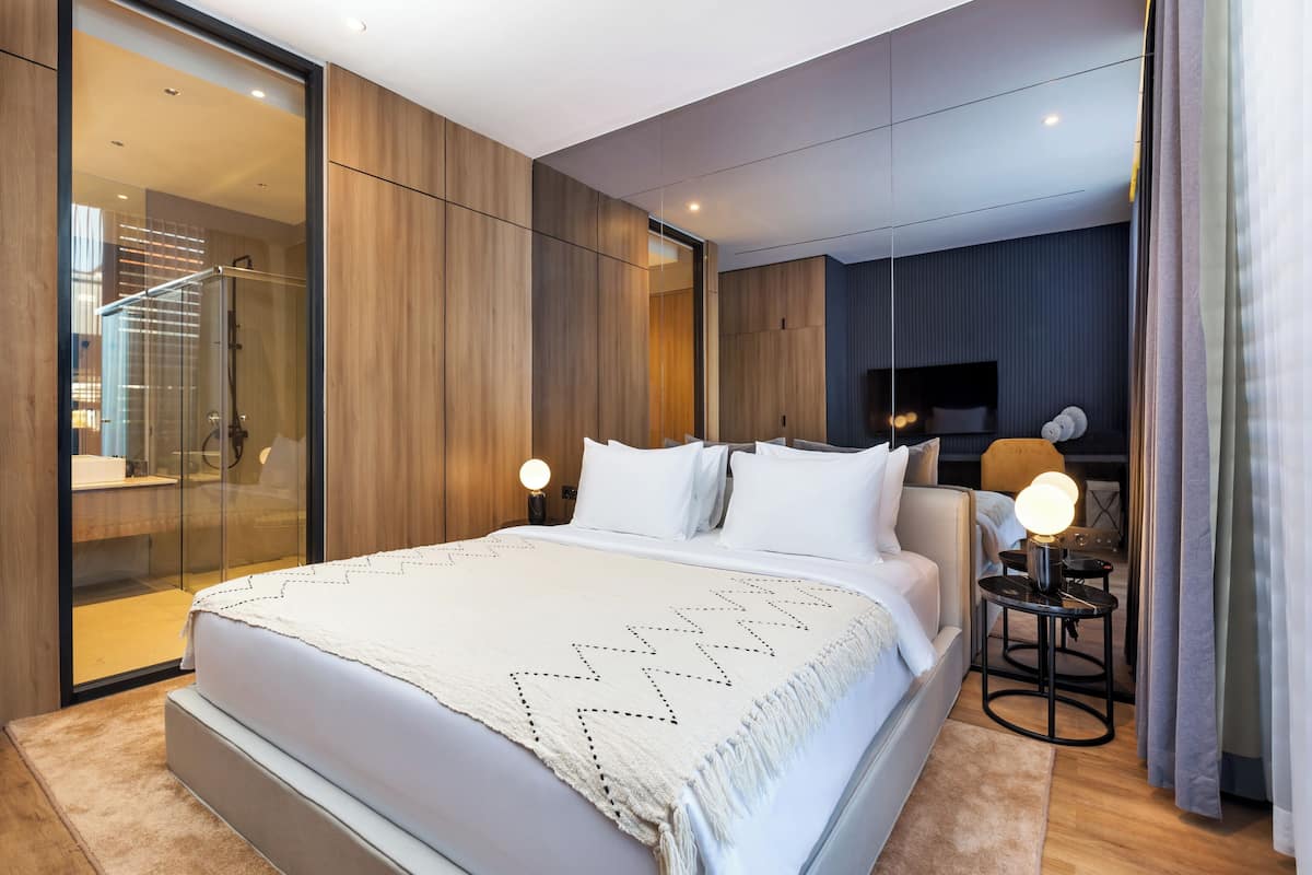 Una habitación de hotel moderna con una cama grande, cabina de ducha de vidrio, mesita de noche con lámpara y televisor sobre un soporte.