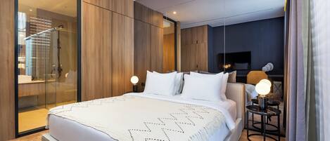 Una habitación de hotel moderna con una cama grande, cabina de ducha de vidrio, mesita de noche con lámpara y televisor sobre un soporte.