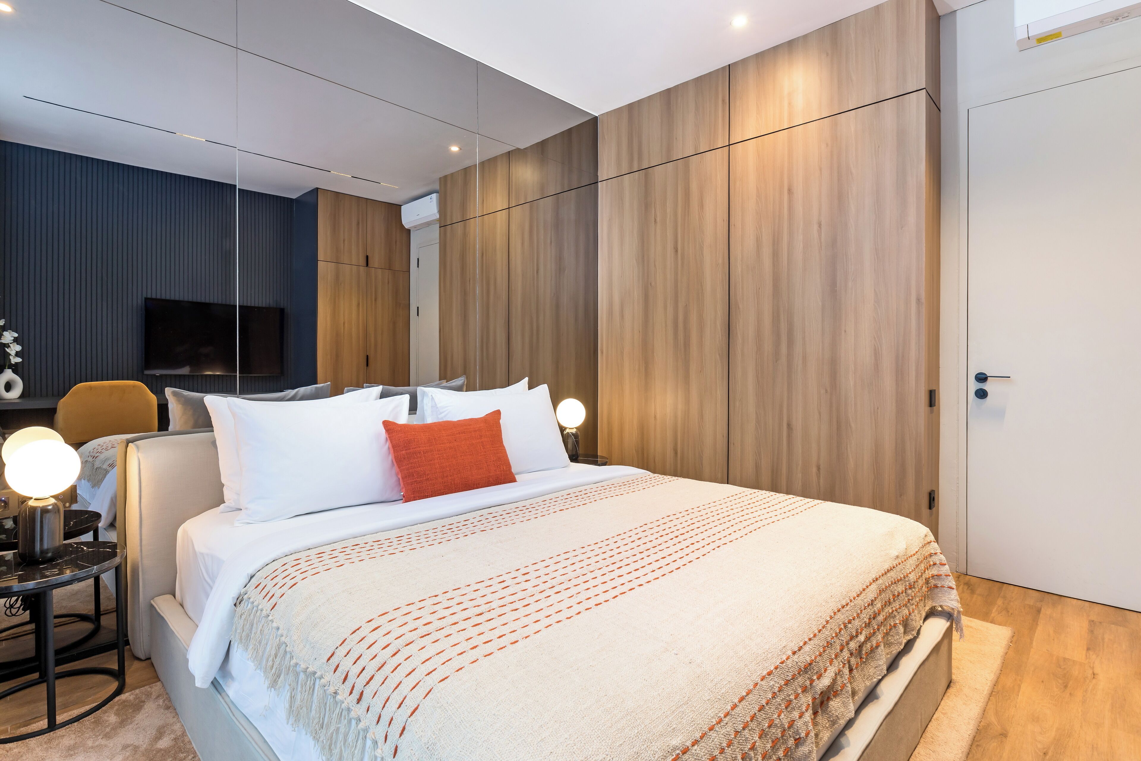 A modern hotel room with a large bed, a flat-screen TV, a desk with a lamp, and a wooden wardrobe.