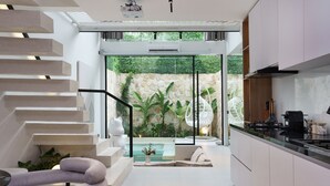 Eine moderne Küche mit einer zentralen Kochinsel, ein Essbereich mit einem weißen Sofa und eine Treppe, die zu einem Balkon mit Poolblick führt.