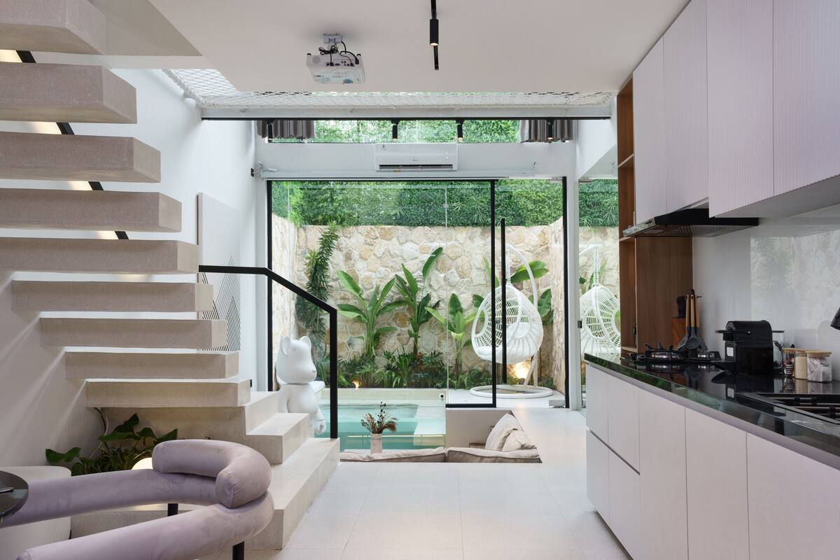 Una cocina moderna con una isla central, un área de comedor con sofá blanco y una escalera que lleva a un balcón con vista a la piscina.