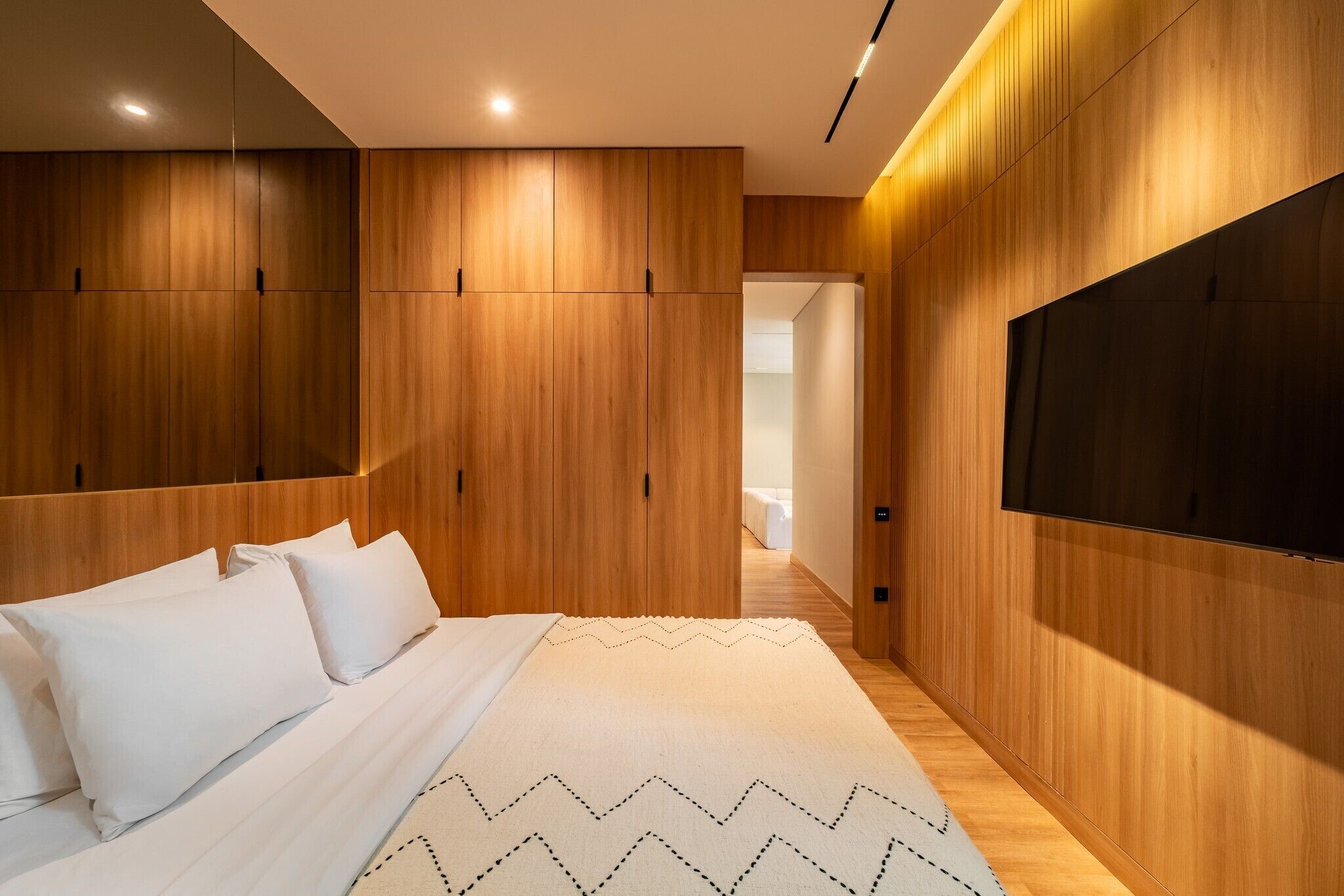 Ein modernes Schlafzimmer mit einem Holzkopfteil, einem flachbildfernseher an der Wand und einem Bett mit weißer Bettwäsche.