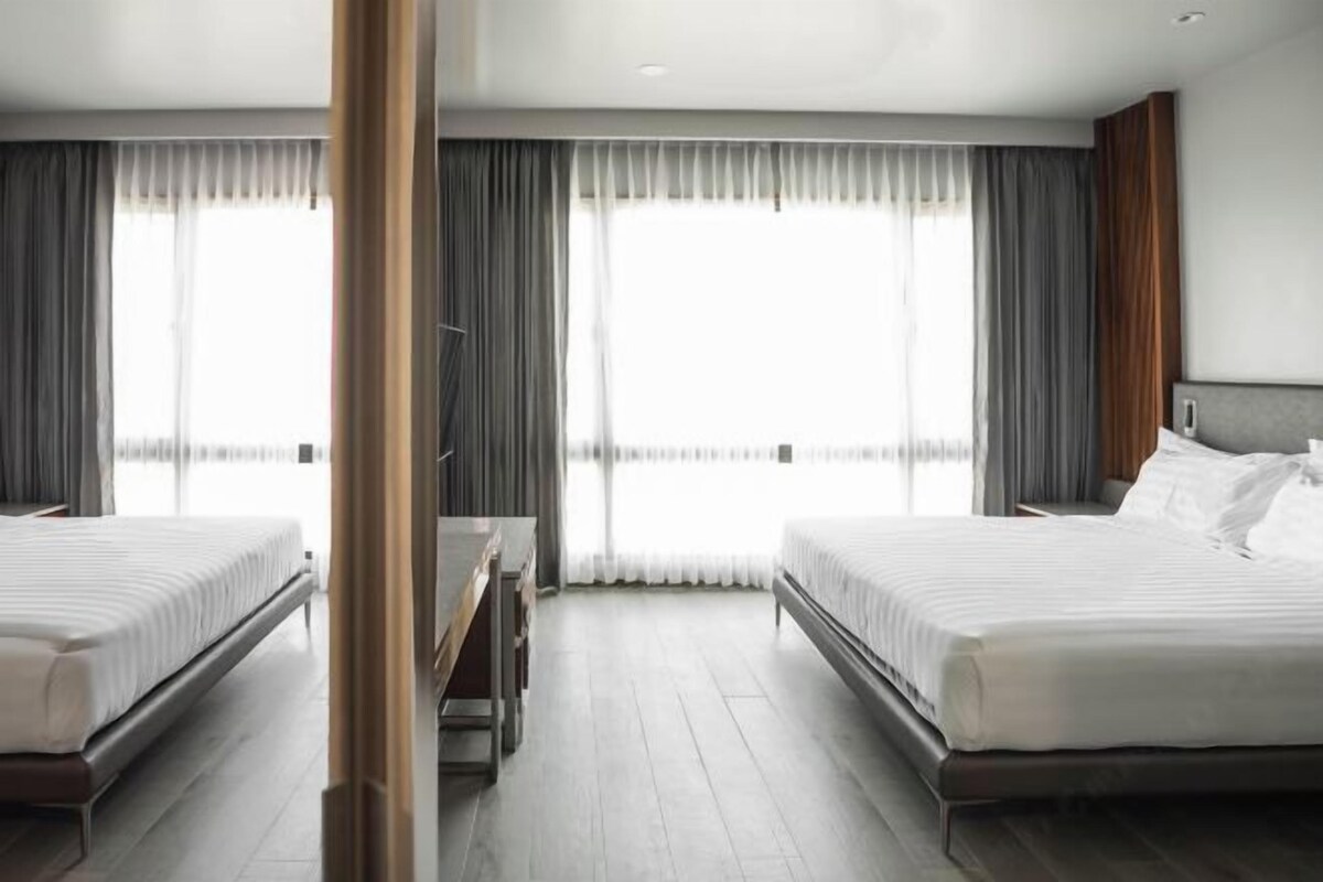 A hotel room with two beds, a desk, and large windows with curtains.