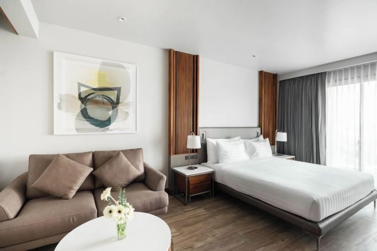 A hotel room with a bed, a sofa, a nightstand, a lamp, and a vase with flowers.
