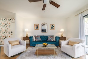 Una sala de estar con un sofá color turquesa, dos sillones blancos, una mesa de centro de vidrio y un ventilador de techo.