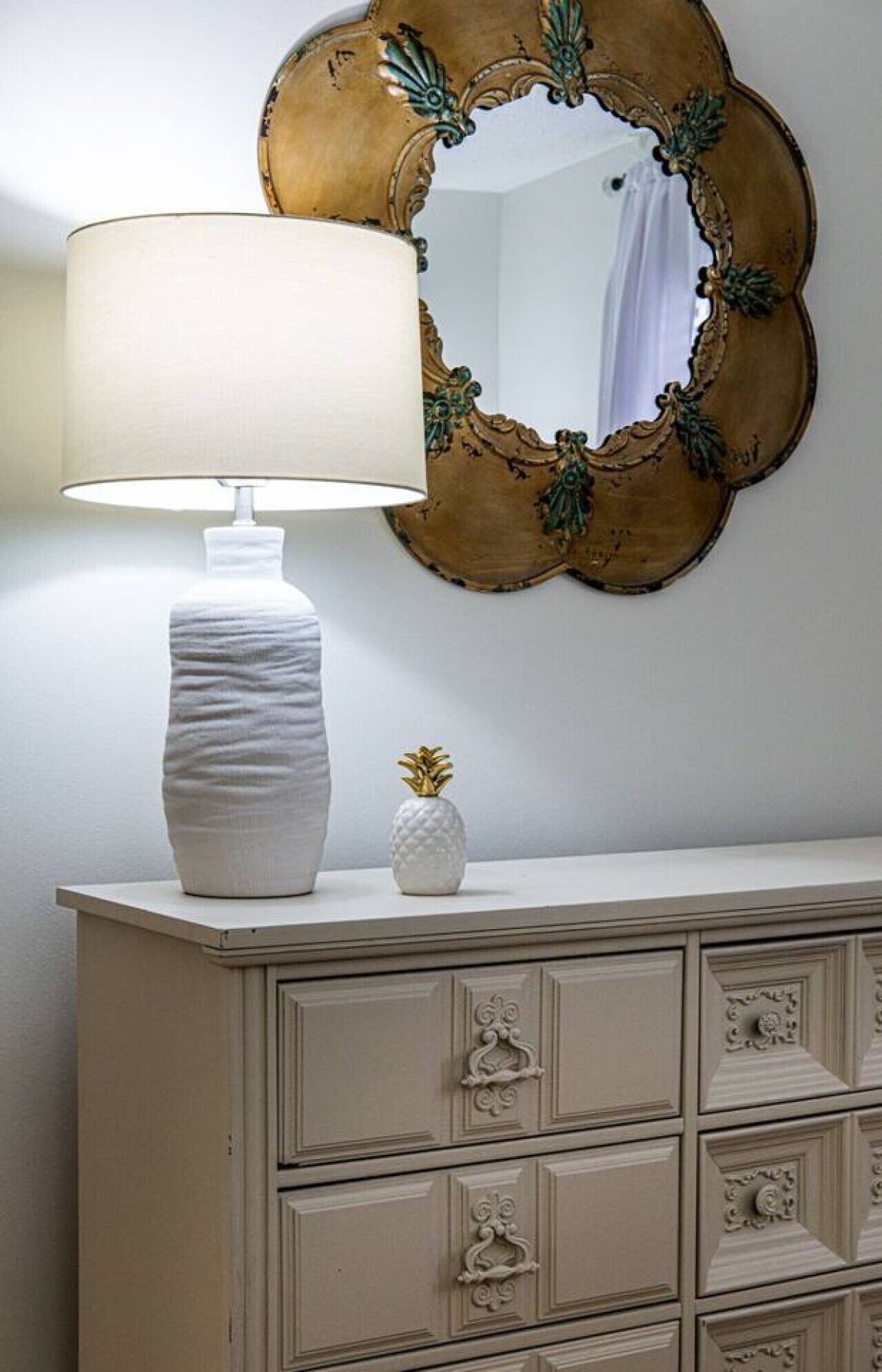 Una lámpara blanca con base texturizada, un espejo decorativo con marco dorado y un pequeño florero con forma de piña sobre una cómoda blanca con varios cajones.