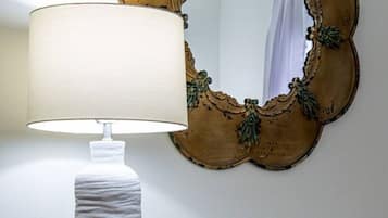Una lámpara blanca con base texturizada, un espejo decorativo con marco dorado y un pequeño florero con forma de piña sobre una cómoda blanca con varios cajones.