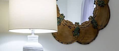 A white lamp with a textured base, a decorative mirror with a gold frame, and a small pineapple-shaped vase on a white dresser with multiple drawers.