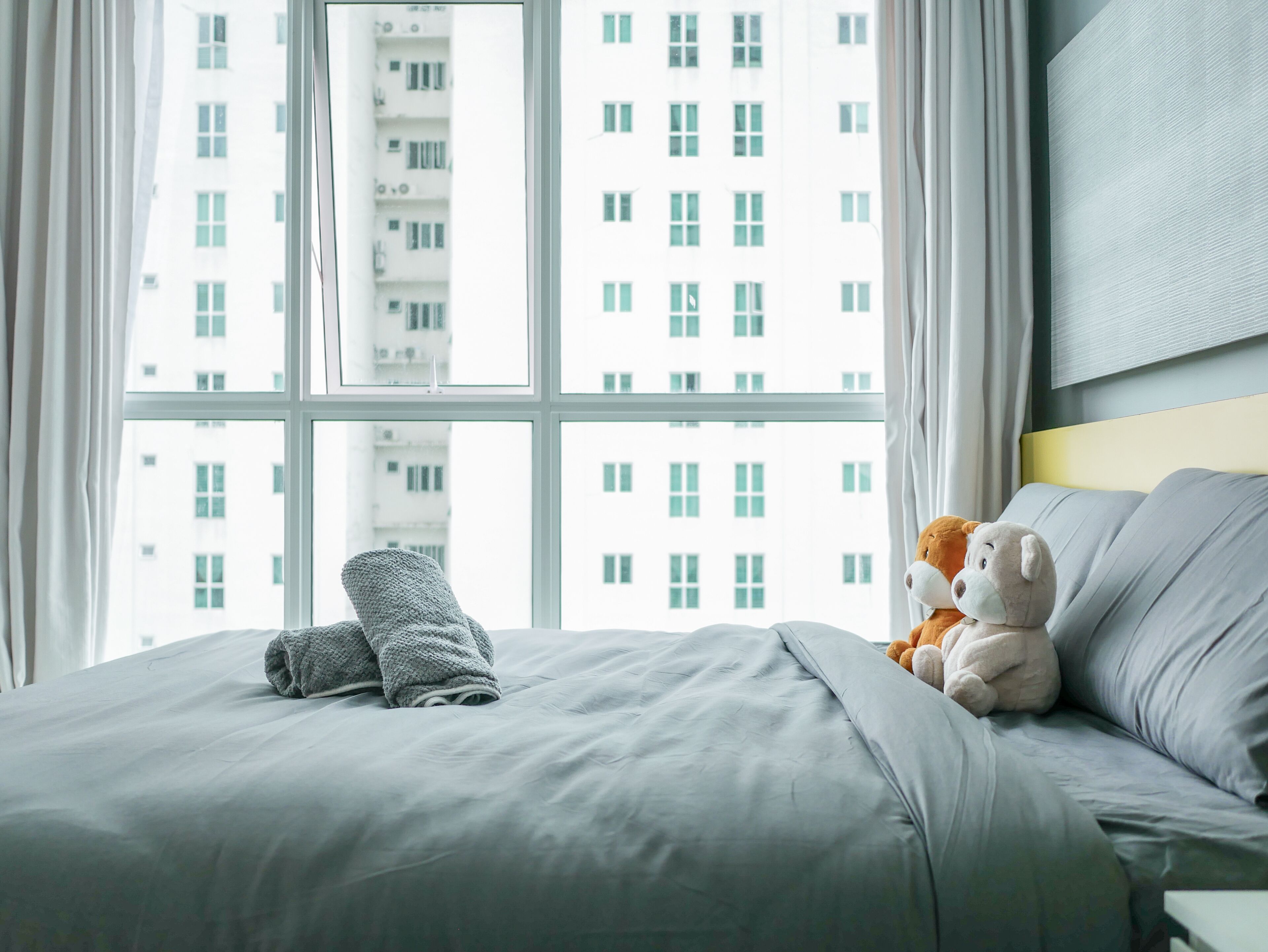 Een bed met een grijze deken en een teddybeer, een raam met uitzicht op een gebouw en een effen muur met een lichtschakelaar.