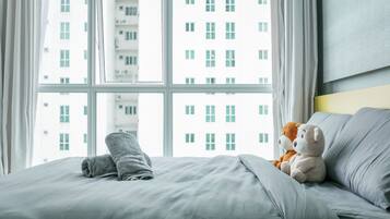 Een bed met een grijze deken en een teddybeer, een raam met uitzicht op een gebouw en een effen muur met een lichtschakelaar.