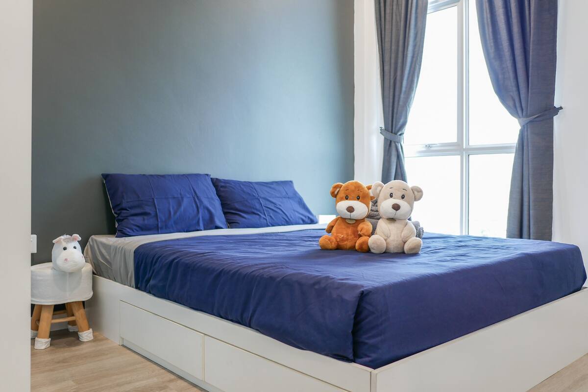 A bedroom with a bed, two stuffed animals, a white chair, and a window with curtains.
