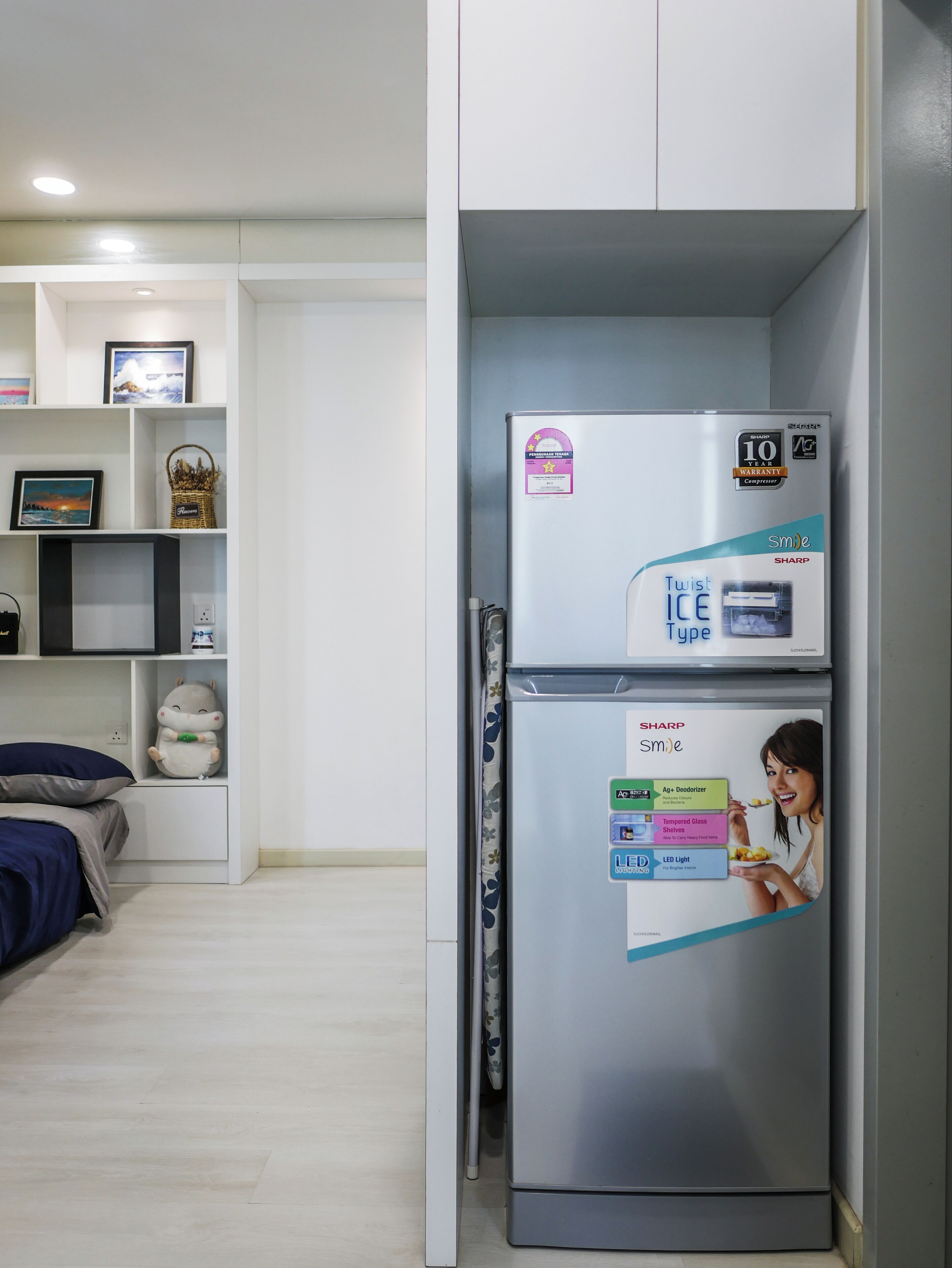 A compact living space with a built-in refrigerator, a bed with a blue pillow, and a small shelf with decorative items.