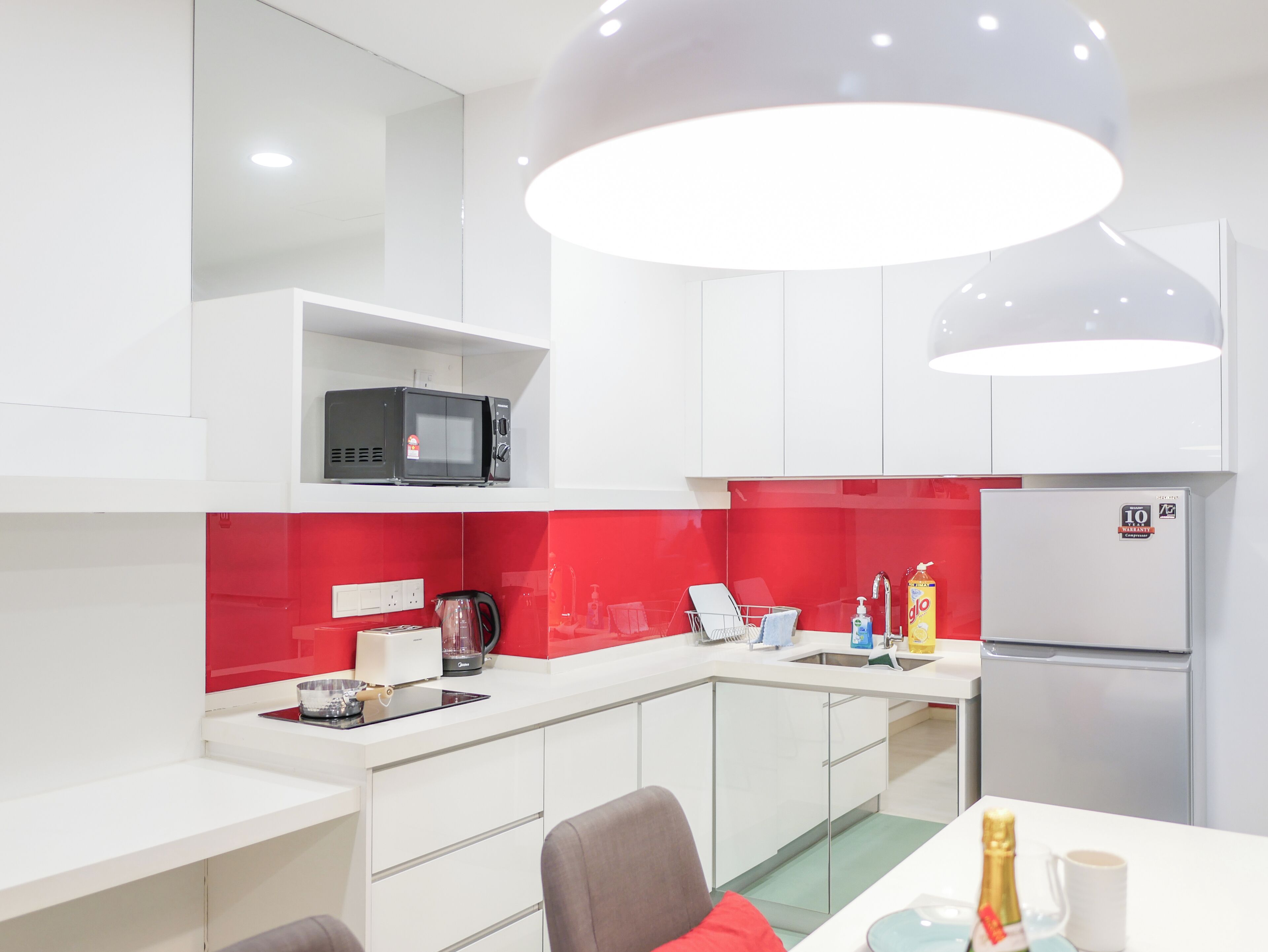 A modern kitchen with white cabinets, a red backsplash, a microwave, a coffee maker, a kettle, and a refrigerator.