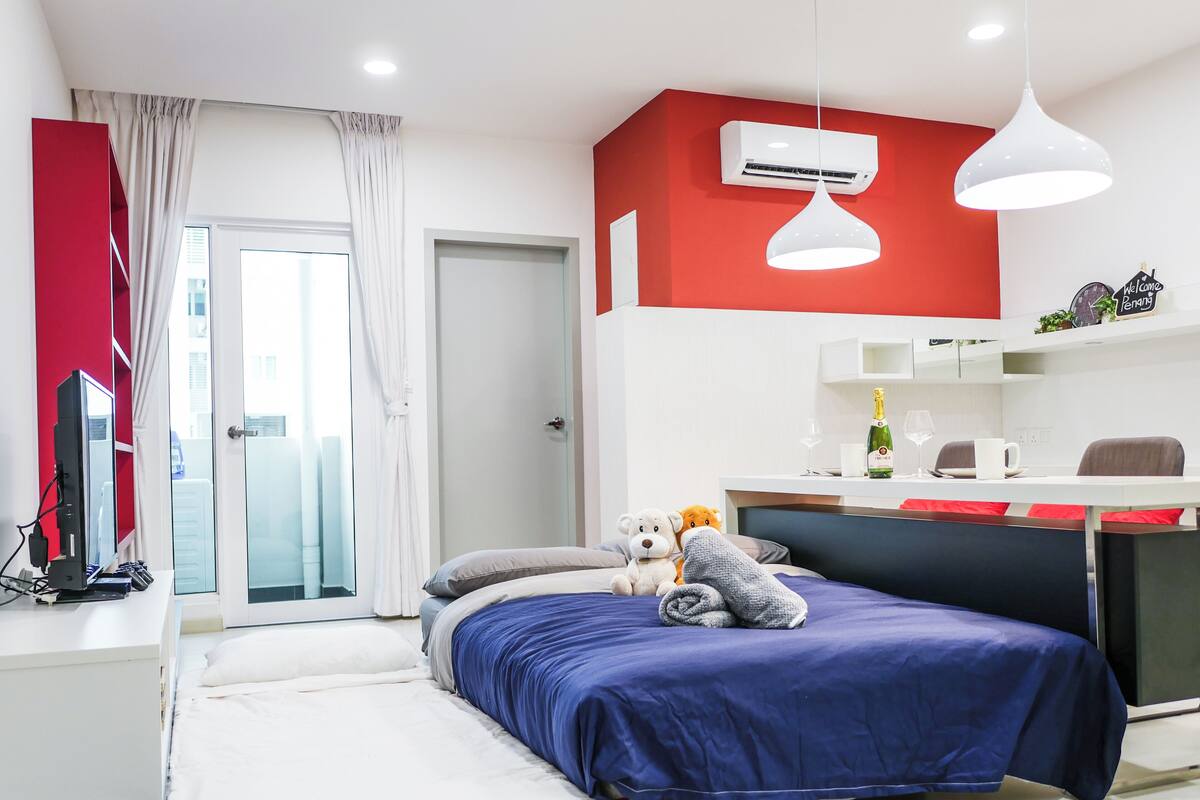 A modern hotel room with a bed, a desk with a computer, a chair, a small table with a bottle and glass, and a red accent wall.