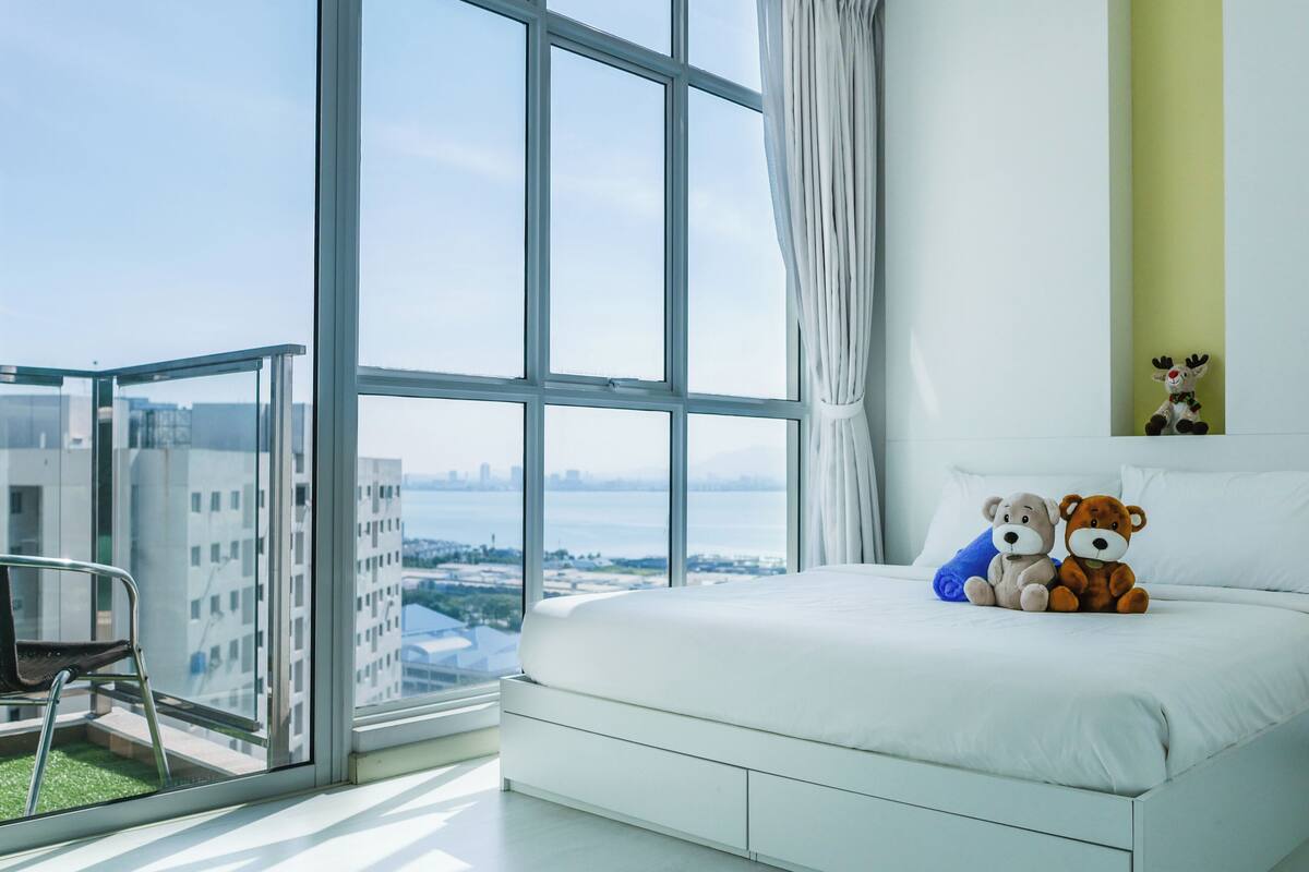 A modern hotel room with a large window, a bed with two plush toys, and a chair by the window.