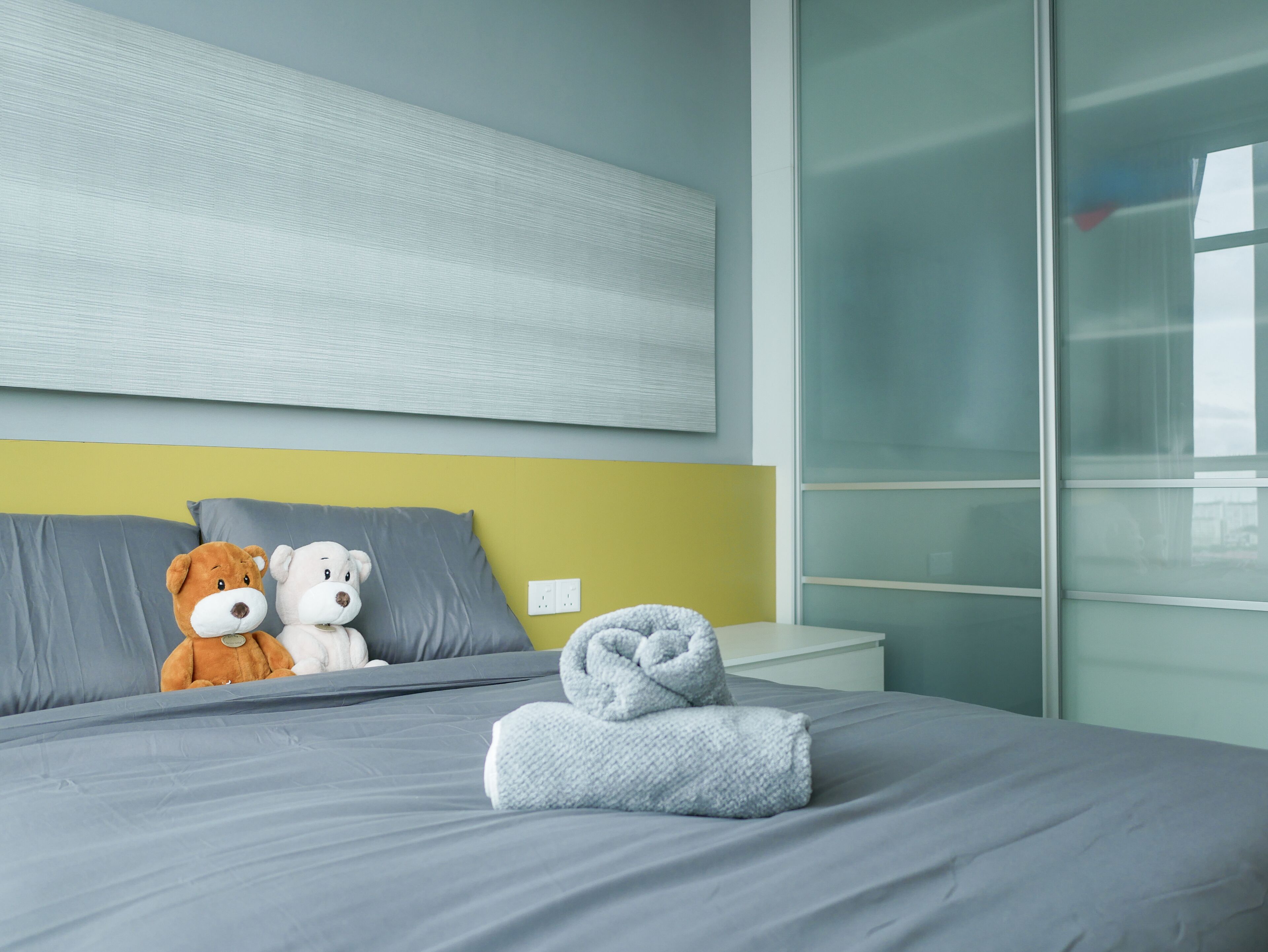 A modern hotel room with a grey bed, a yellow accent wall, and a large window.
