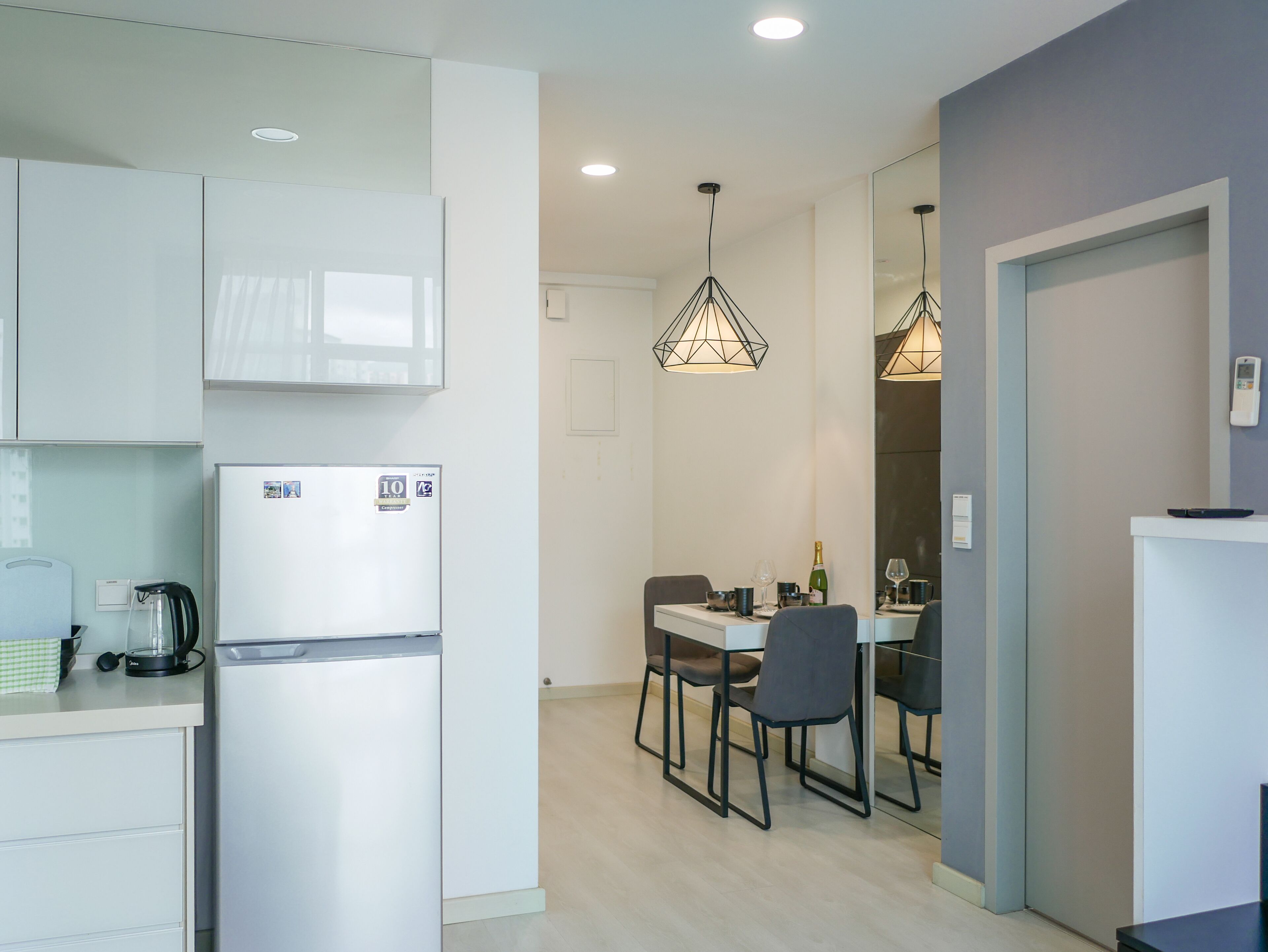 A modern kitchen with white cabinets, a built-in microwave, and a refrigerator. There is a dining area with a table and chairs, and pendant lights above.