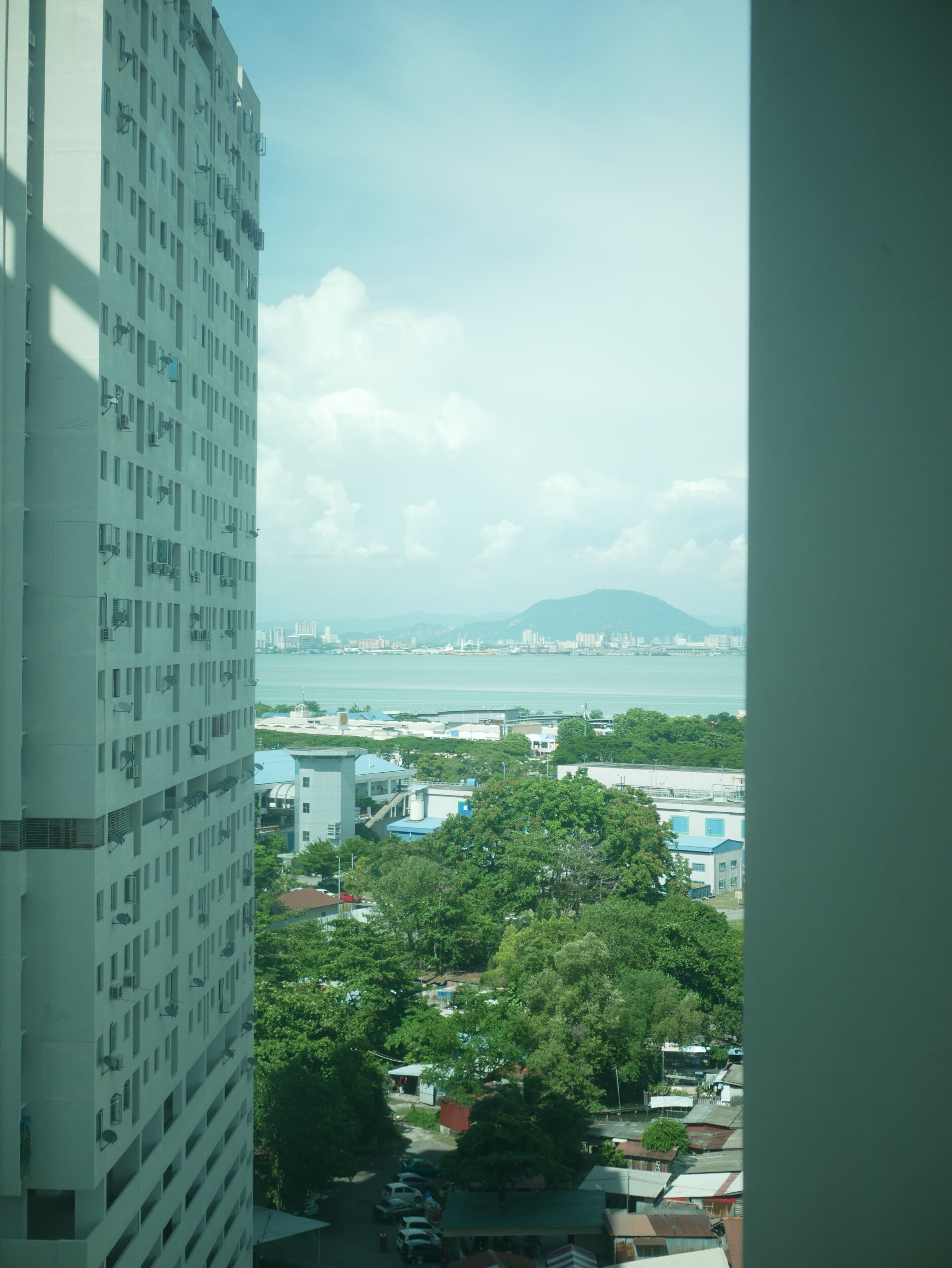 A high-rise building with multiple floors and balconies, overlooking a coastal area with buildings and greenery.