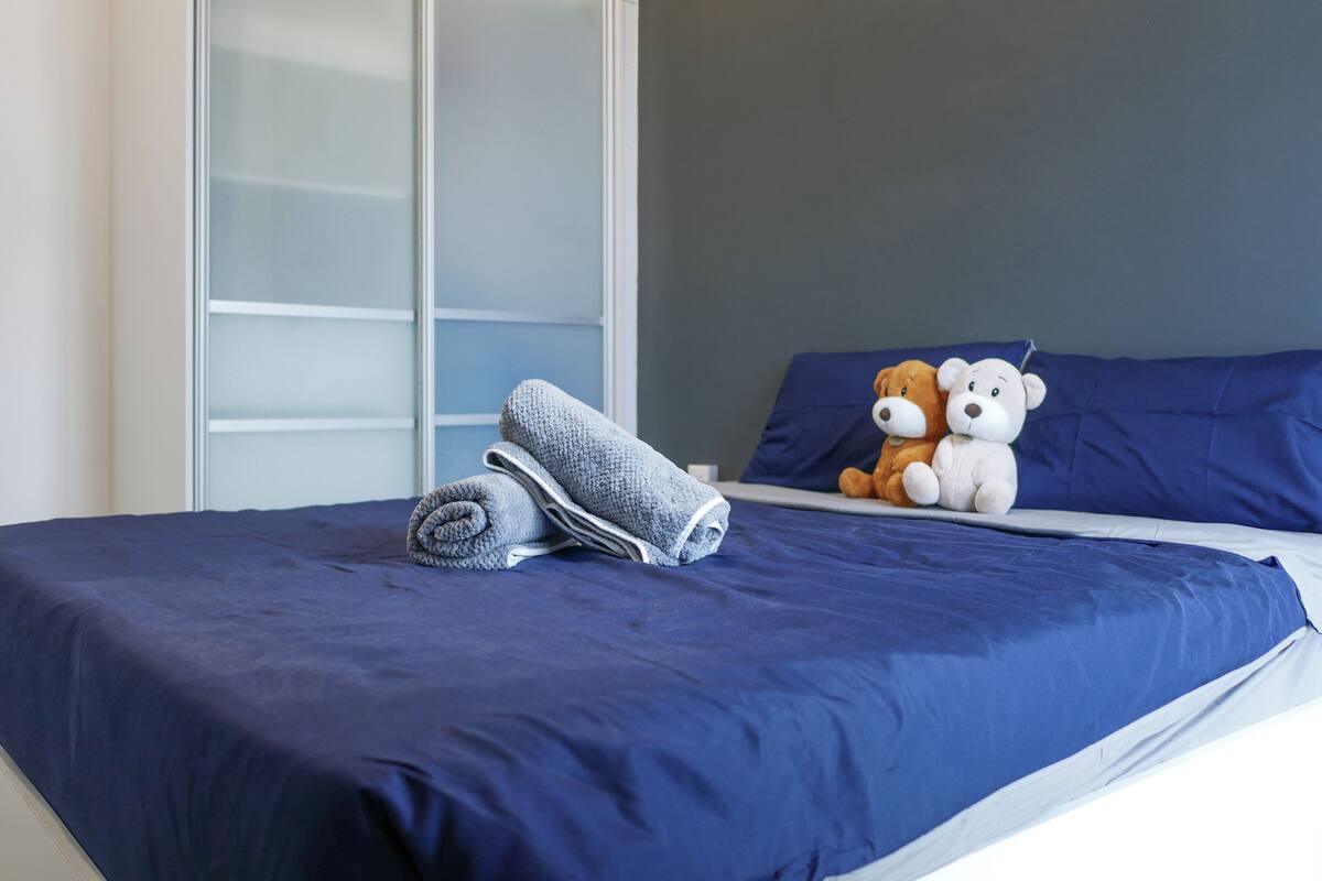 A neatly made bed with a blue bedspread and pillows, a pair of folded towels, and two stuffed teddy bears on the bed.
