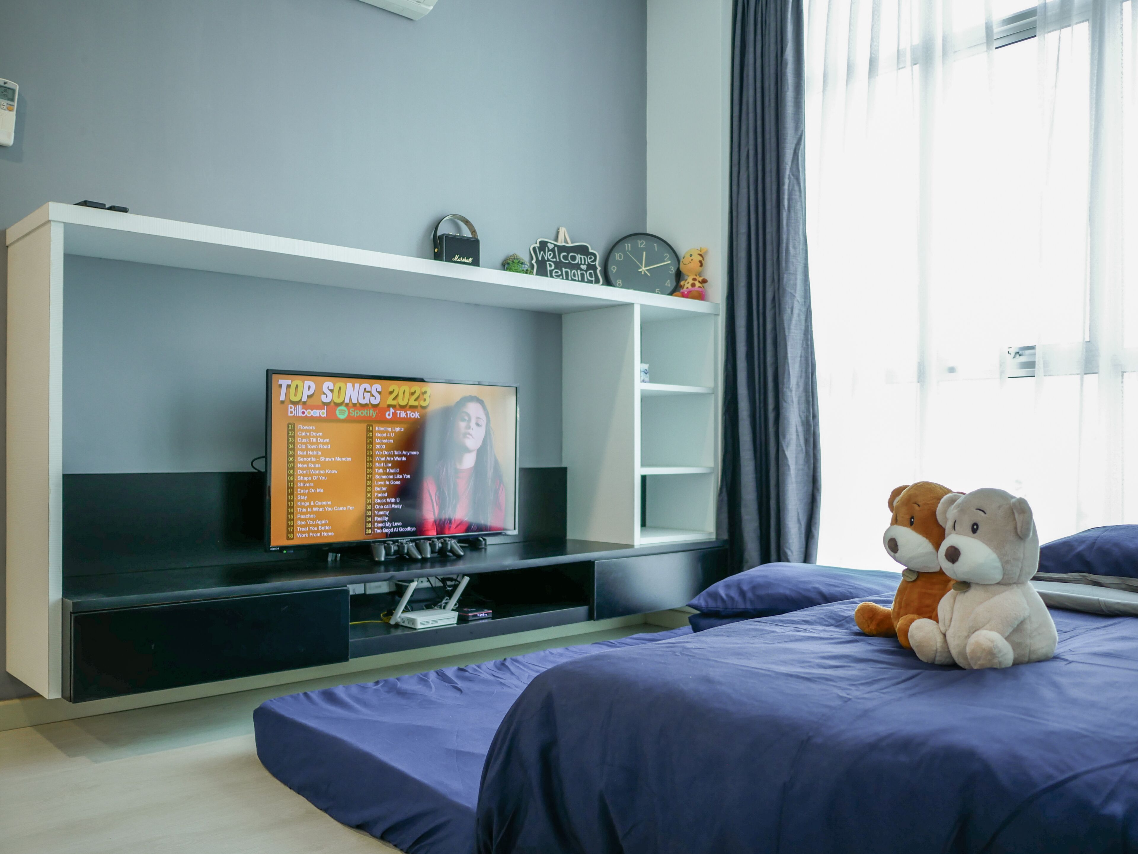 A modern bedroom with a TV displaying a list of top songs, a shelf with decorative items, a clock, and plush toys on the bed.