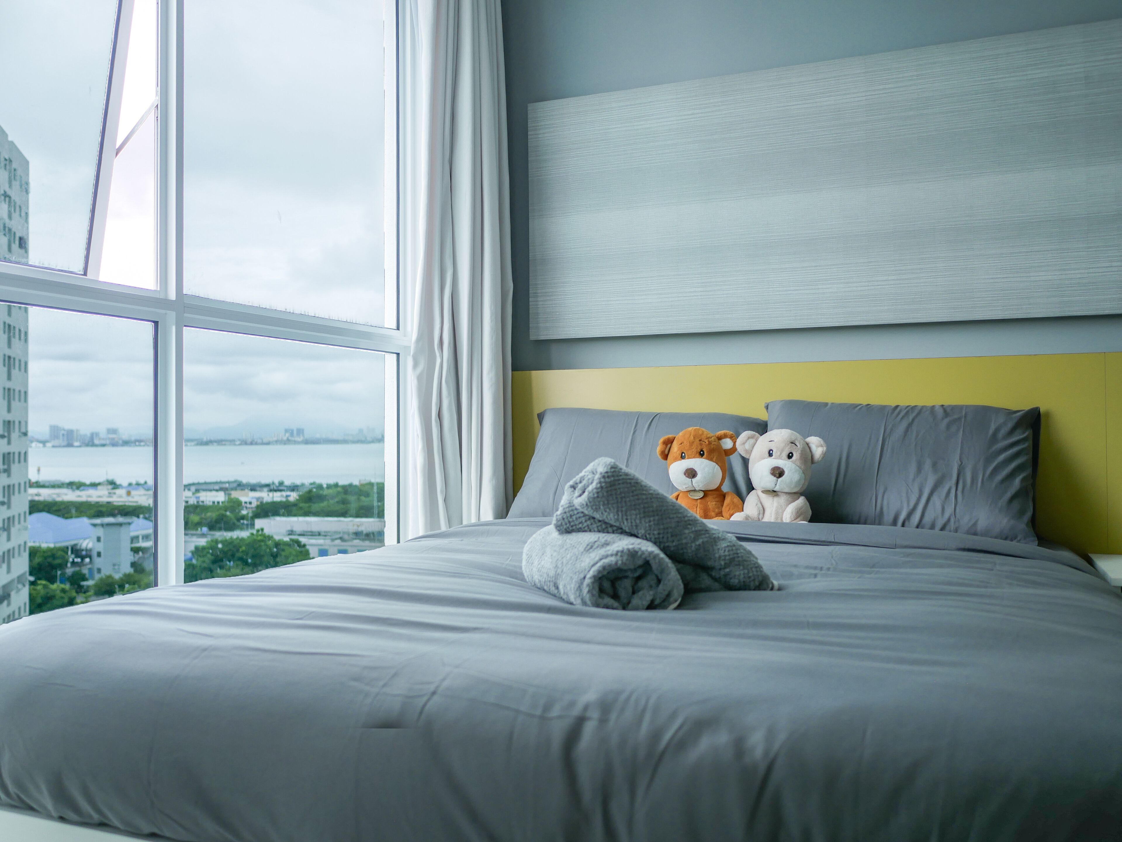 Ein modernes Hotelzimmer mit einem großen Bett, Blick auf eine Stadtlandschaft und zwei weiche Plüschtiere auf dem Bett.