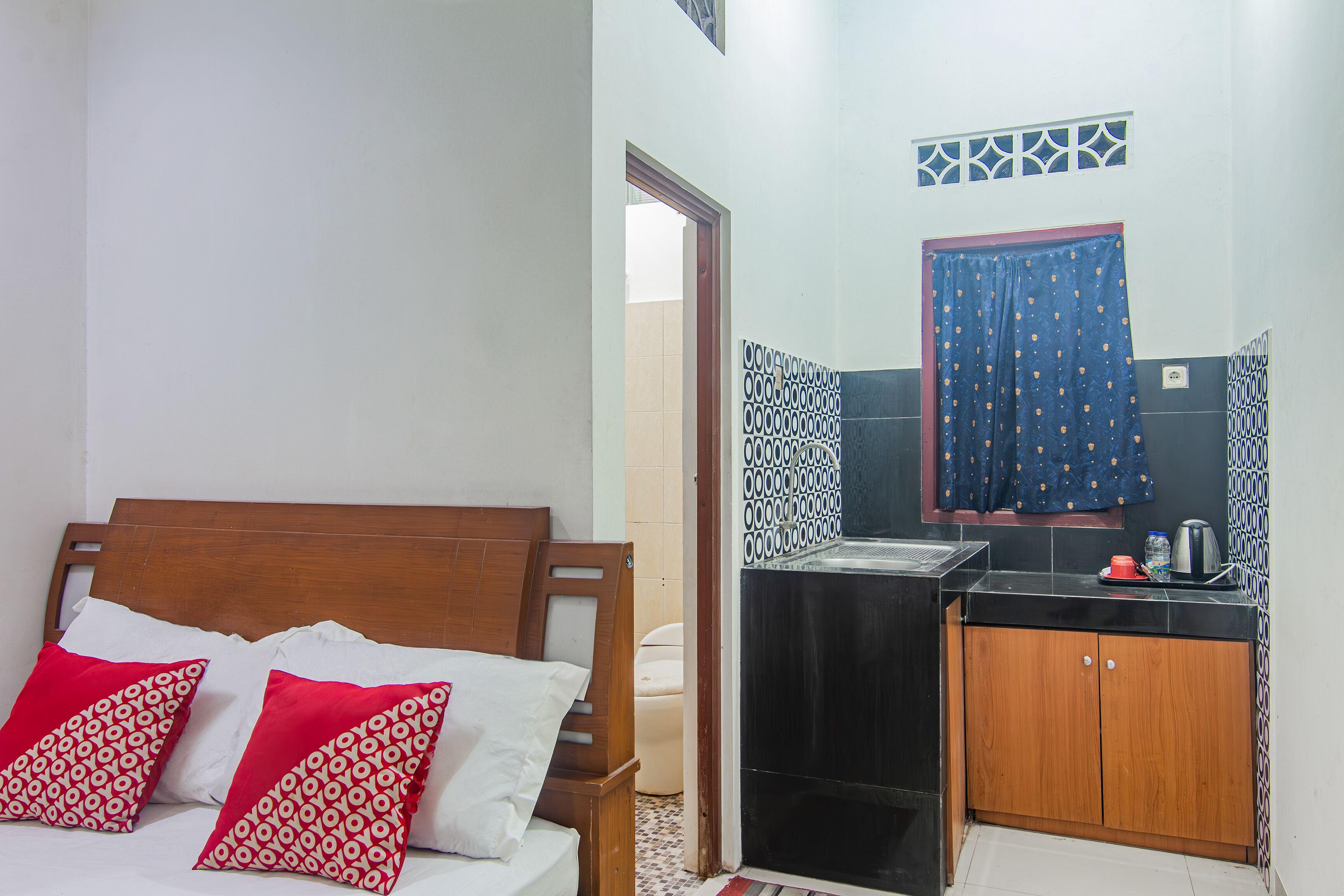 A bedroom with a wooden bed, red and white pillows, a small kitchenette with a sink and cabinets, and a bathroom visible through an open door.