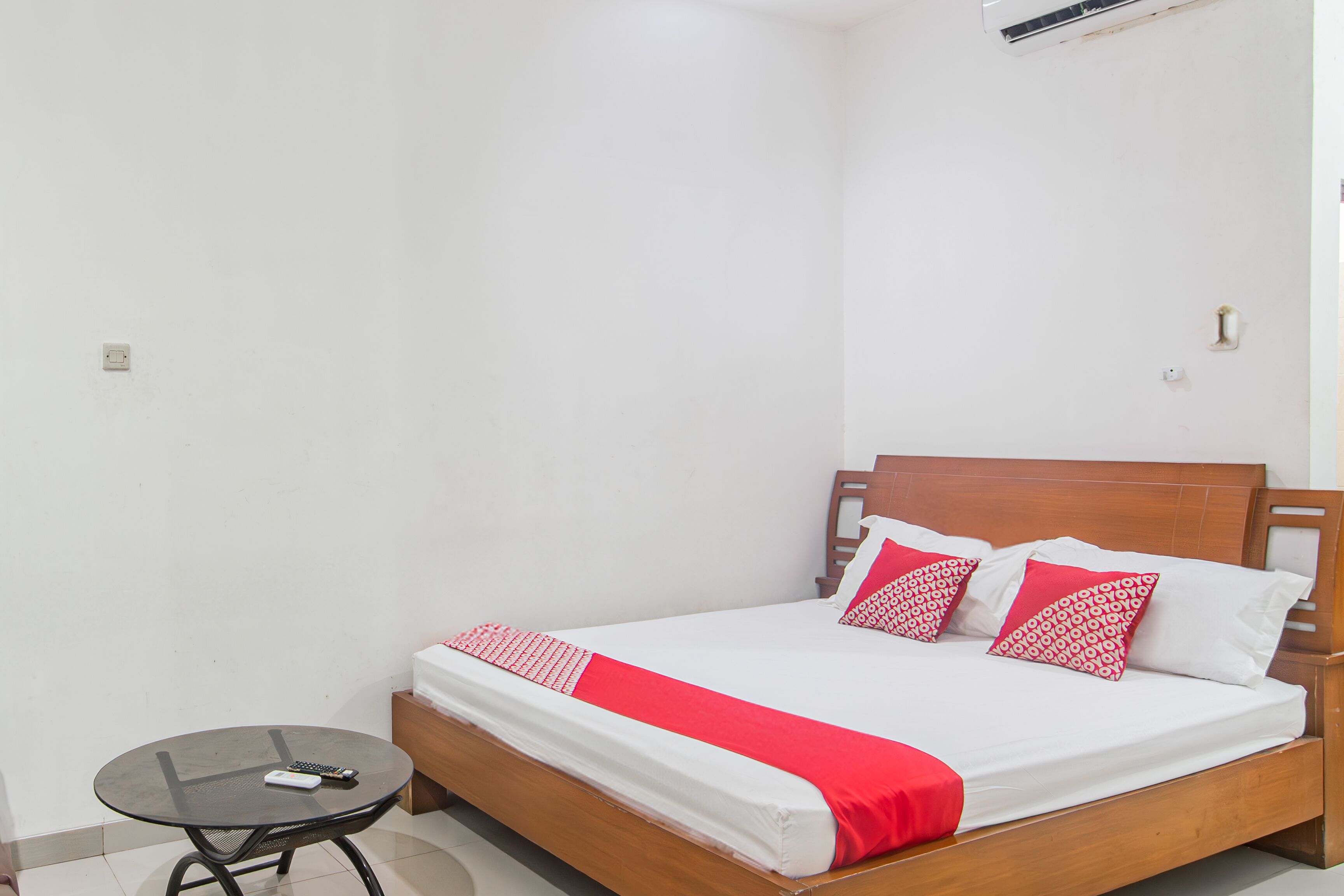 A neatly made bed with a wooden headboard, a small round table with a remote control, and a red and white bedspread.