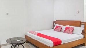 Een netjes opgemaakt bed met een houten hoofdbord, een klein rond tafeltje met een afstandsbediening en een rood-witte dekbedsprei.