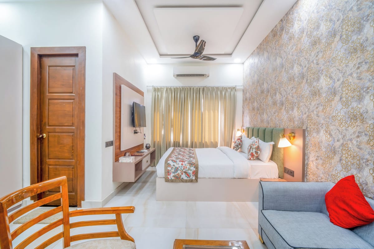 A hotel room with a bed, a sofa, a chair, a TV, a bedside table, and a ceiling fan.