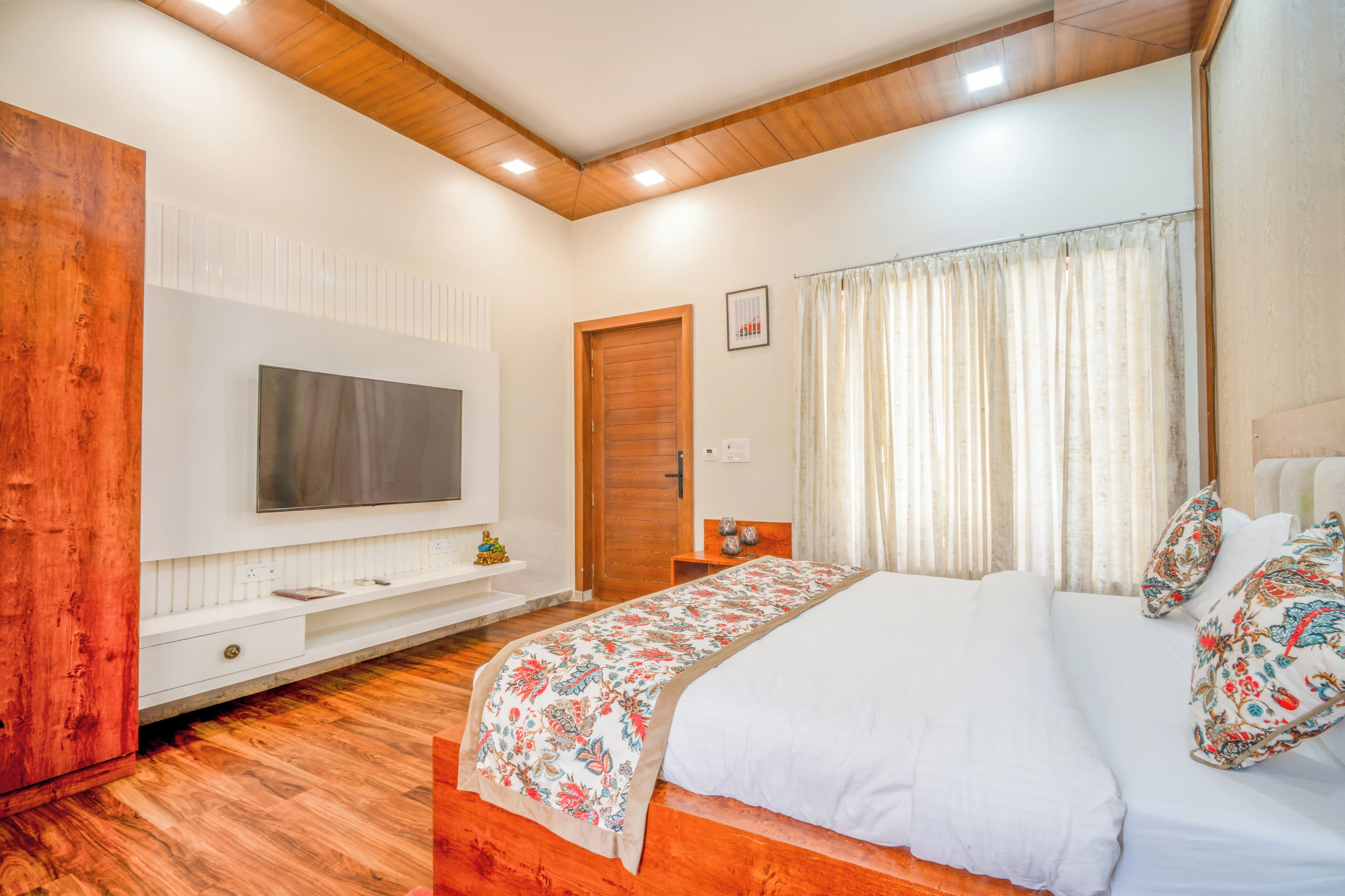 A bedroom with a wooden ceiling, a large bed with floral bedding, a TV mounted on the wall, and a wooden wardrobe.
