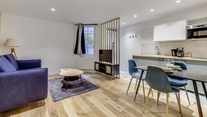 Ein modernes Wohnzimmer mit einer Couch, einem Couchtisch und einem Fernseher. Es gibt einen Essbereich mit Stühlen und einem Tisch. Die Küche verfügt über Schränke und eine Arbeitsplatte.