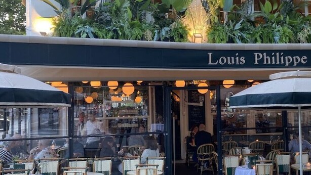 Ein Straßencafé mit Tischen und Stühlen, einem Schild mit der Aufschrift "Louis Philippe" und einer Glasveranda mit Pflanzen.