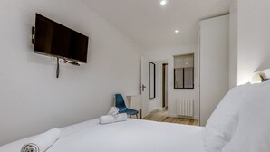 Ein modernes Hotelzimmer mit einem großen Bett, einem flachbildfernseher an der Wand, einem blauen Sessel und einem Fenster mit Blick auf den Flur.
