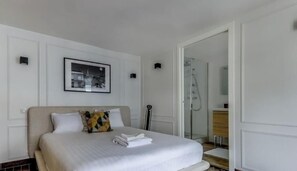 Habitación de hotel con cama, un cuadro en la pared, cabina de ducha de vidrio y mesita de noche de madera.
