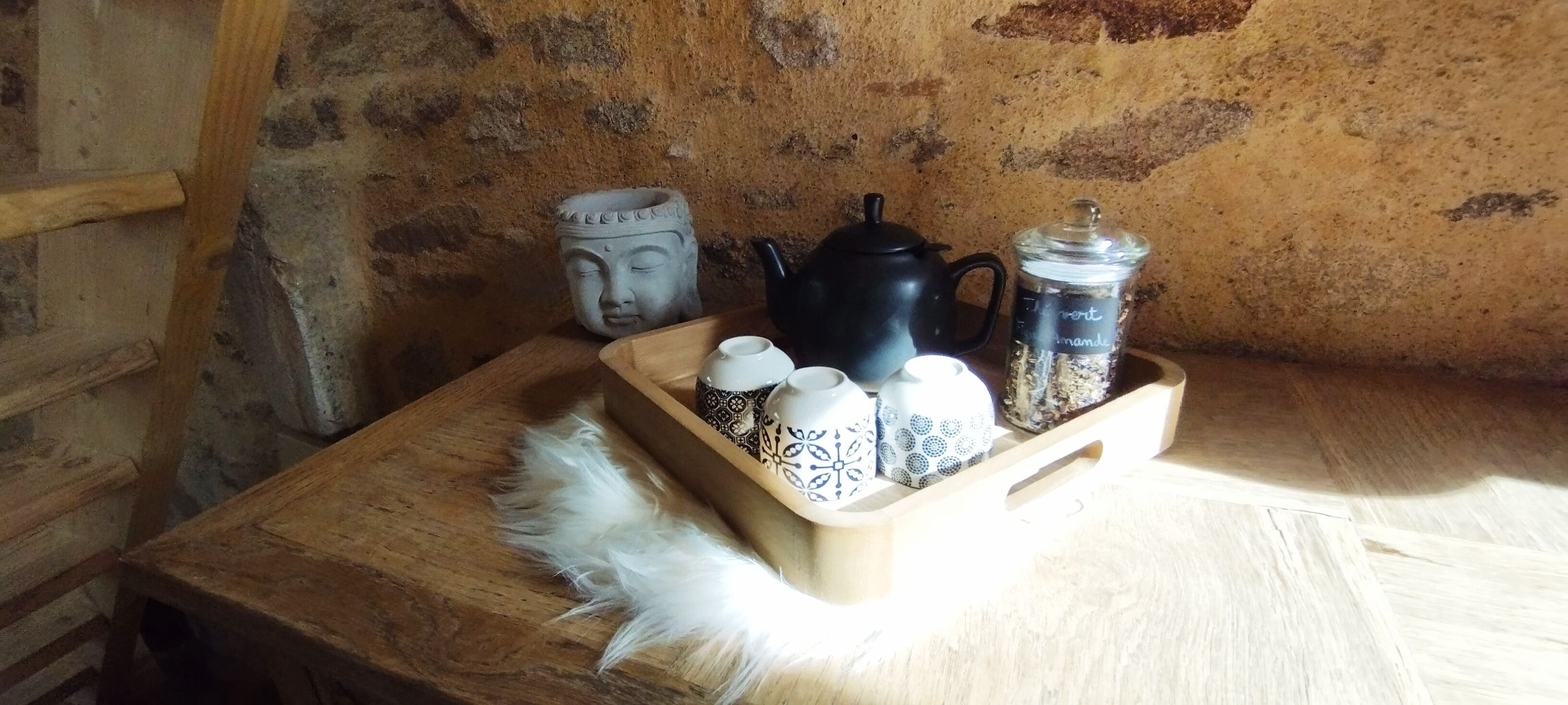 A wooden table with a tray holding a black teapot, a ceramic mug, a jar with a label, and several patterned cups. A white feather is also visible on the table.
