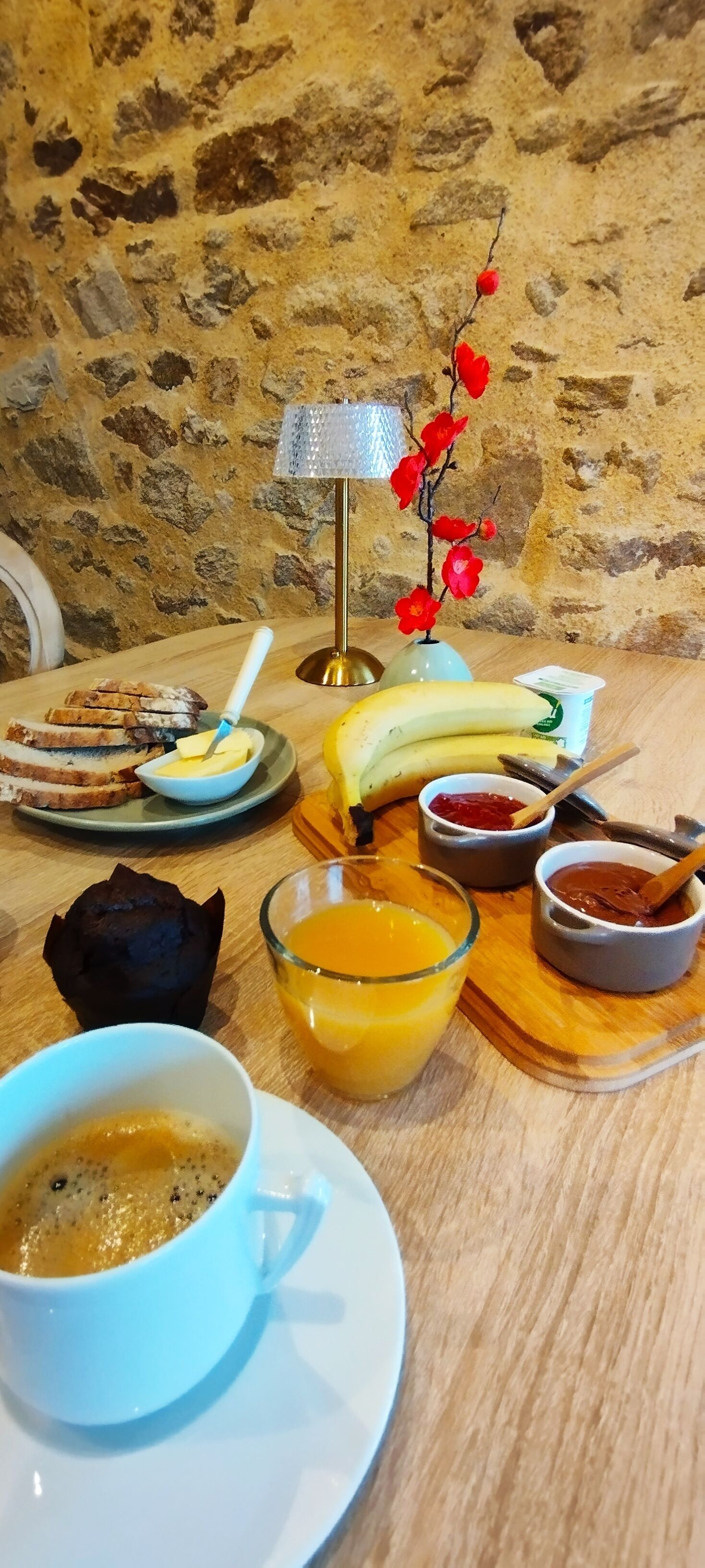 A breakfast setting with bread, banana, orange juice, coffee, and condiments on a wooden table against a stone wall.