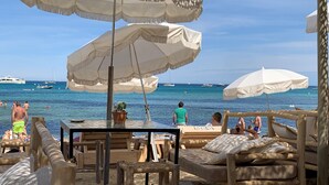 A beachside setting with wooden lounge chairs and tables under white umbrellas, overlooking a calm sea with boats. - Charming stone house with airco and a large pool (Lorgues)