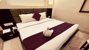 Ein ordentlich bezogenes Bett mit einer weiß-violetten Tagesdecke, einem Kopfteil mit dunkler Verkleidung und einem kleinen weißen Dekoartikel auf dem Bett.