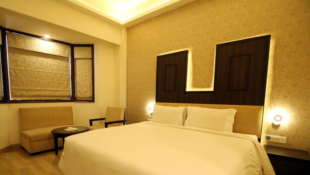 A hotel room with a large bed, a sofa, a small table, and a chair. There are two wall-mounted lamps and a window with curtains. - Hotel Forest Avenue (Dehradun)
