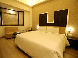 A hotel room with a large bed, a sofa, a small table, and a chair. There are two wall-mounted lamps and a window with curtains. - Hotel Forest Avenue (Dehradun)
