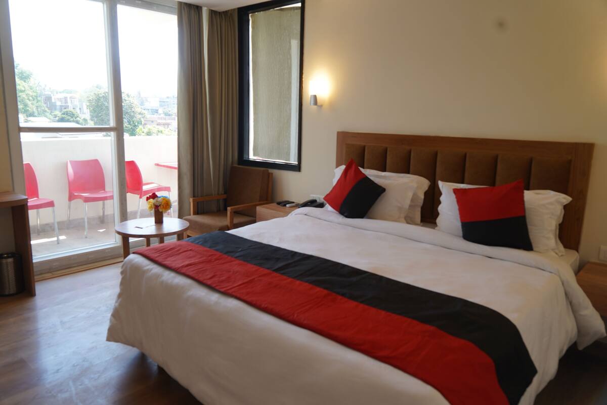 A hotel room with a large bed, a wooden headboard, a small table with a vase of flowers, and a balcony with red chairs.