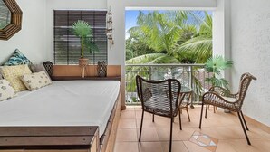 A bedroom with a bed, a balcony with wicker chairs, and a view of palm trees.
