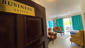 A hotel room with a sign indicating 'BUSINESS SUITES'. The room has a large window with curtains, a seating area with chairs, and a small table.