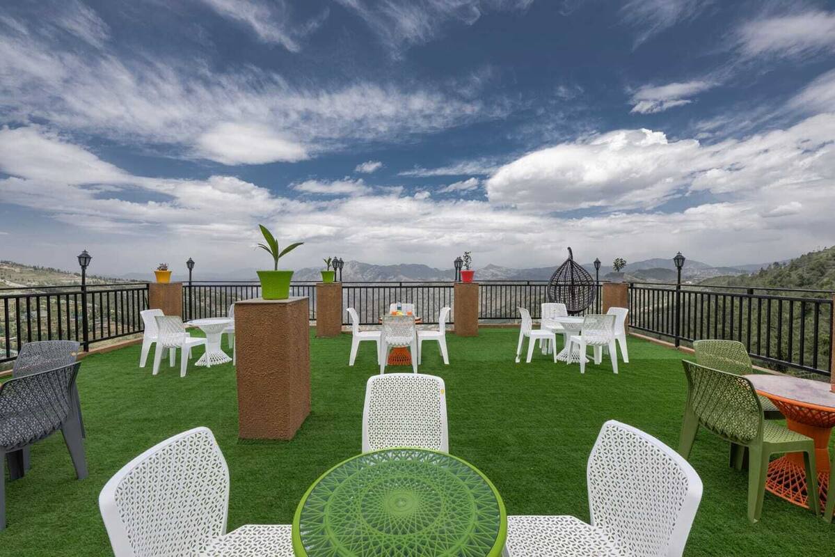 Una terraza al aire libre con sillas y mesas blancas, rodeada por una barandilla negra, con vistas a un paisaje montañoso.