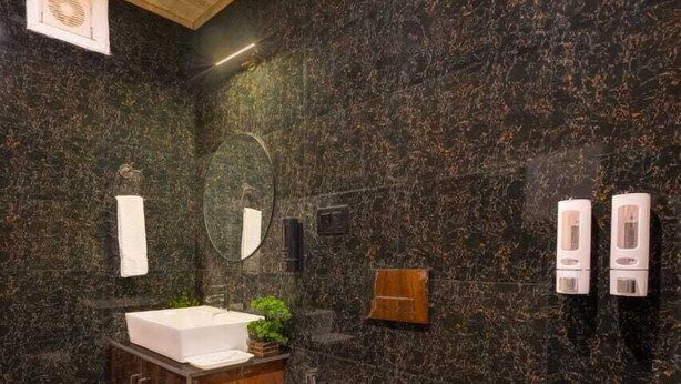 Un baño moderno con techo de madera, paredes de mármol oscuro, un lavabo blanco, inodoro y zona de ducha.