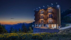 Un edificio de varios pisos con balcones, rodeado por un paisaje montañoso y un campo de flores amarillas.